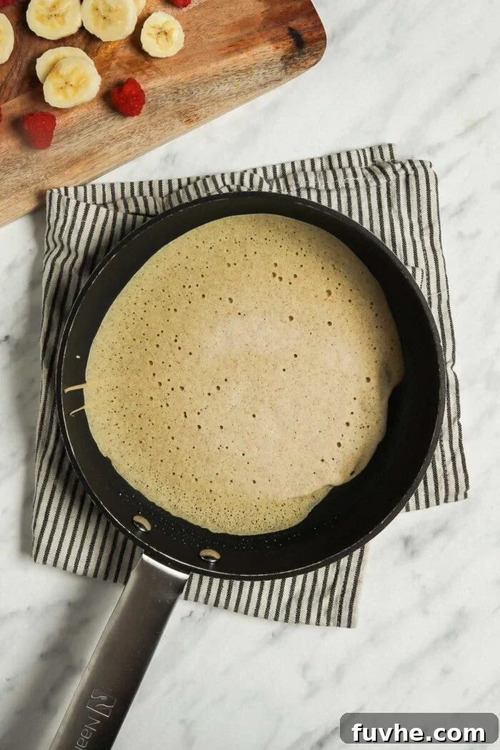 A thin vegan buckwheat crepe cooking in a hot skillet, with finely chopped strawberries and banana pieces visible on a cutting board next to the pan, ready to be used as fillings.