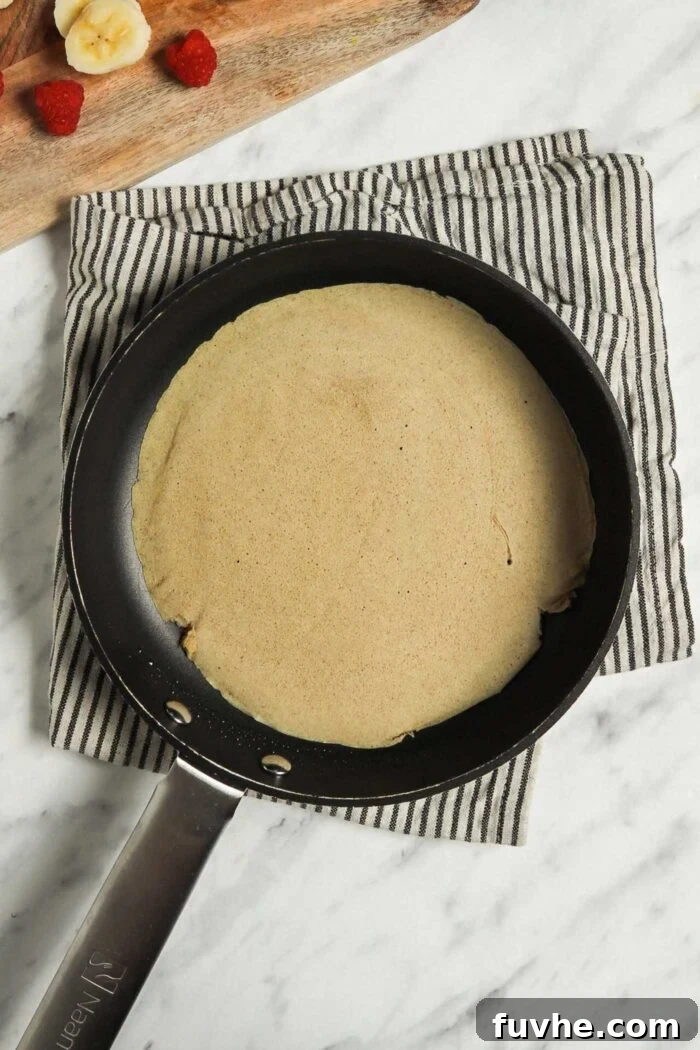 A perfectly cooked vegan buckwheat crepe resting in a skillet after being flipped, alongside a wooden cutting board laden with fresh strawberries and sliced bananas, highlighting the readiness for assembly.