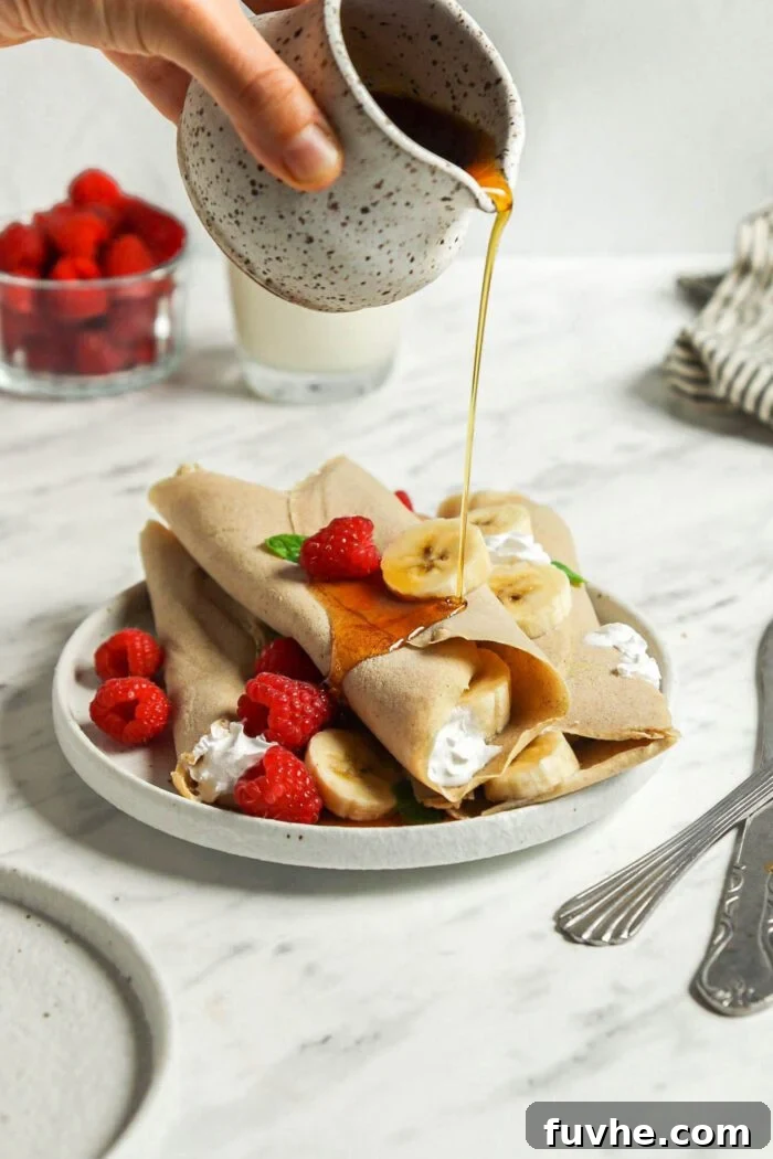 A hand gently pours golden maple syrup from a small ceramic pitcher over a tempting stack of vegan buckwheat crepes, artfully arranged on a plate and filled with vibrant strawberries and ripe banana slices, enhancing their appeal.