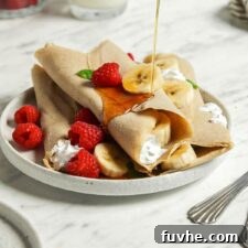 A stack of golden brown vegan buckwheat crepes filled with fresh strawberries and banana, drizzled with maple syrup, presented beautifully on a plate.