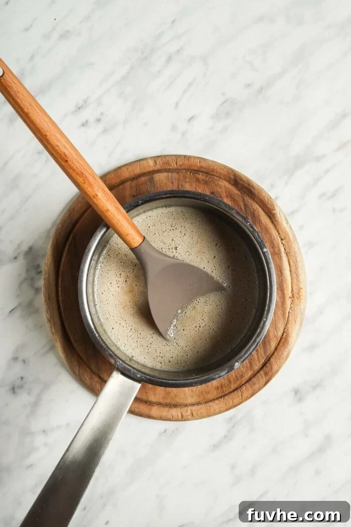 Liquid caramel mixture cooking in a saucepan with a wooden spoon, showing its developing golden hue.
