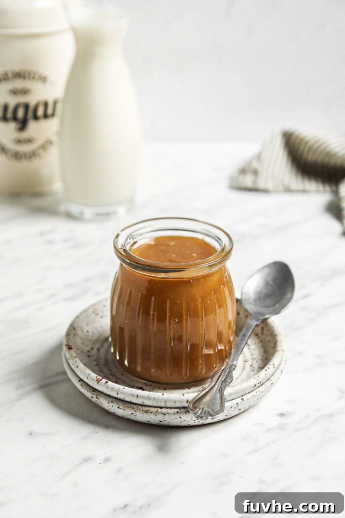 Small jar of homemade caramel sauce on a plate with a spoon beside it, ready to be enjoyed.