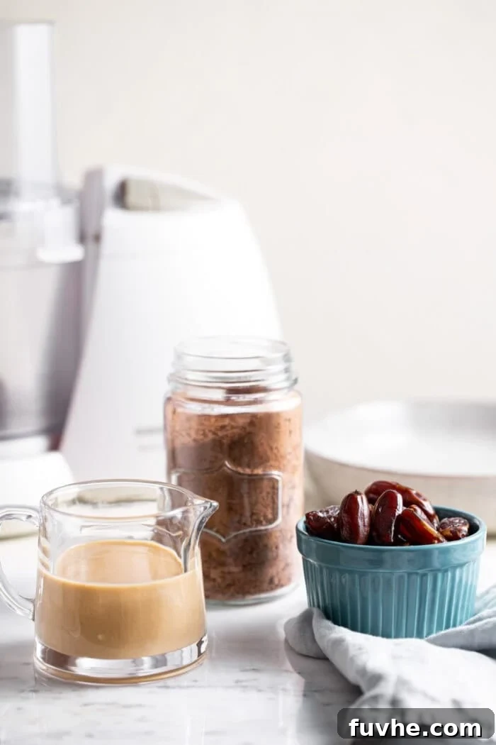 Containers of tahini, cacao powder and dates sitting in front of a food processor.