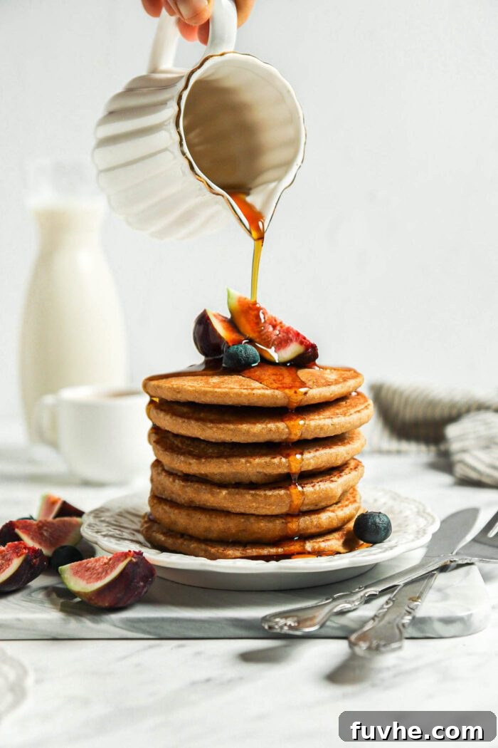 Hand pouring maple syrup from a small container over a stack of healthy oatmeal pancakes.