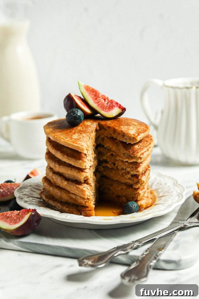 Stack of thick vegan oat flour pancakes topped with blueberries and figs on a small plate.The stack has a slice taken from it so you can see the inside texture of the pancakes.