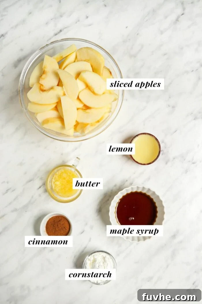 Apples, lemon, butter, maple syrup, cinnamon and cornstarch in bowls on a marble counter.