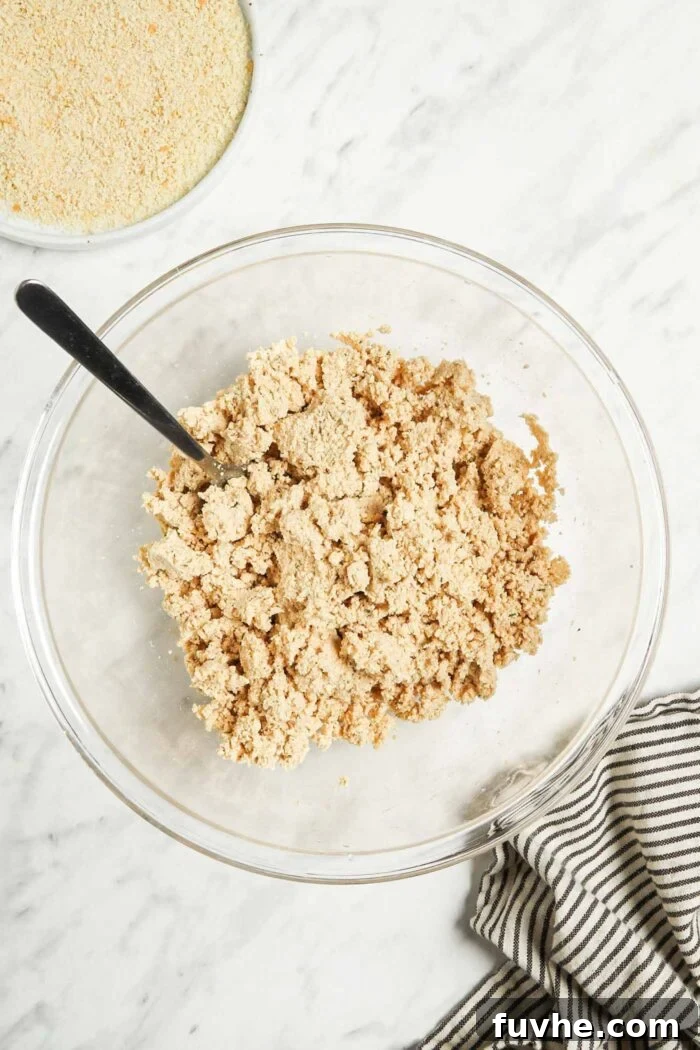 Crispy Baked Tofu Bites 5 A glass mixing bowl holds the crumbly tofu mixture after chickpea flour and breadcrumbs have been added, being mixed with a spoon to achieve a cohesive texture.