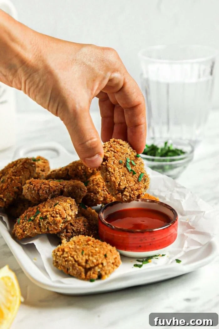 Crispy Baked Tofu Bites 8 A hand gently dips a golden-baked tofu nugget into a small, inviting bowl of vibrant red ketchup, showcasing its crispy texture.