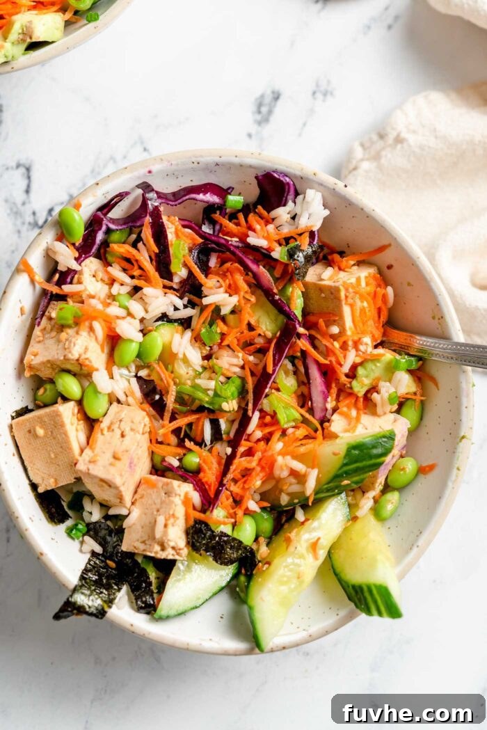 Rice, tofu, cucumber, edamame, cabbage, carrot, sesame seeds and nori strips mixed up in a bowl with a fork in it.