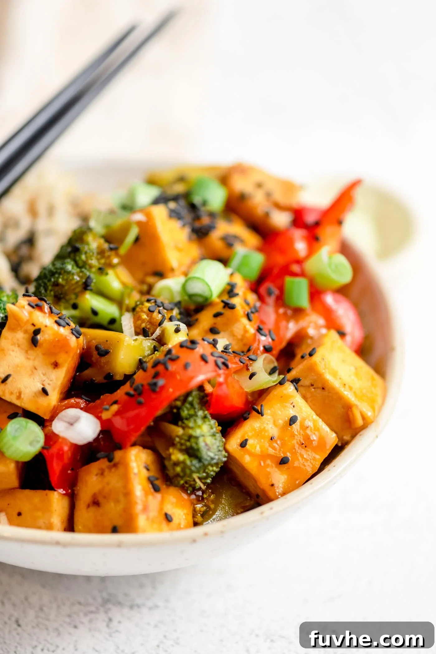 Pair of chopsticks rests on a bowl of stir fried vegetables and tofu in orange sauce.