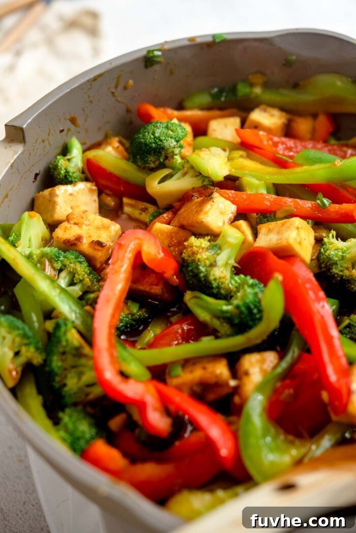 Cubed tofu and stir fried vegetables cooking in a stick orange sauce in a pan.