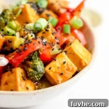 Pair of chopsticks rests on a bowl of stir fried vegetables and tofu in orange sauce.