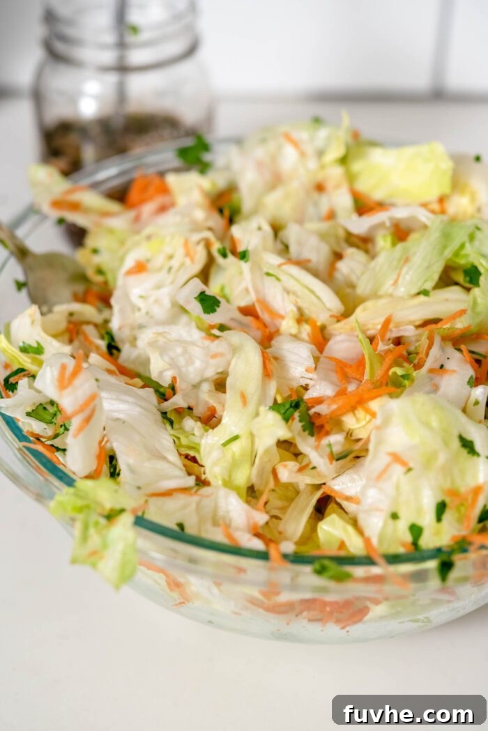 A person combining and mixing chopped lettuce (or cabbage), grated carrot, thinly sliced scallions, and fresh cilantro in a large mixing bowl.