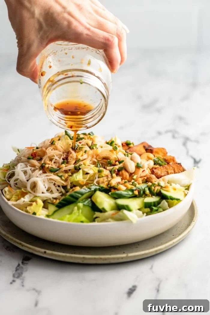 A hand pouring a generous amount of vibrant salad dressing over a vermicelli noodle salad, beautifully garnished with peanuts and cucumber slices.