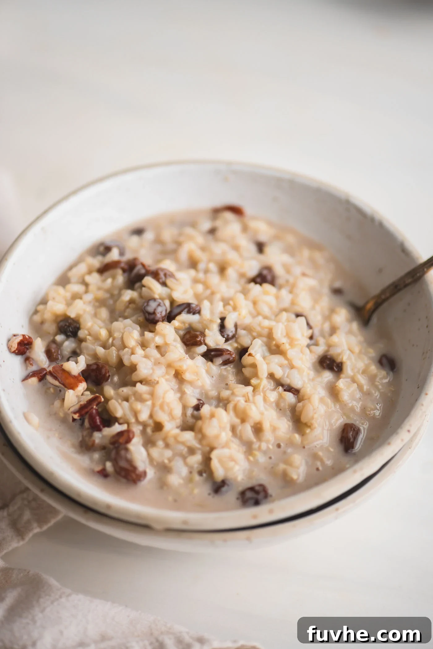 Bowl of brown rice pudding with raisins and pecans. A spoon rests in the bowl.