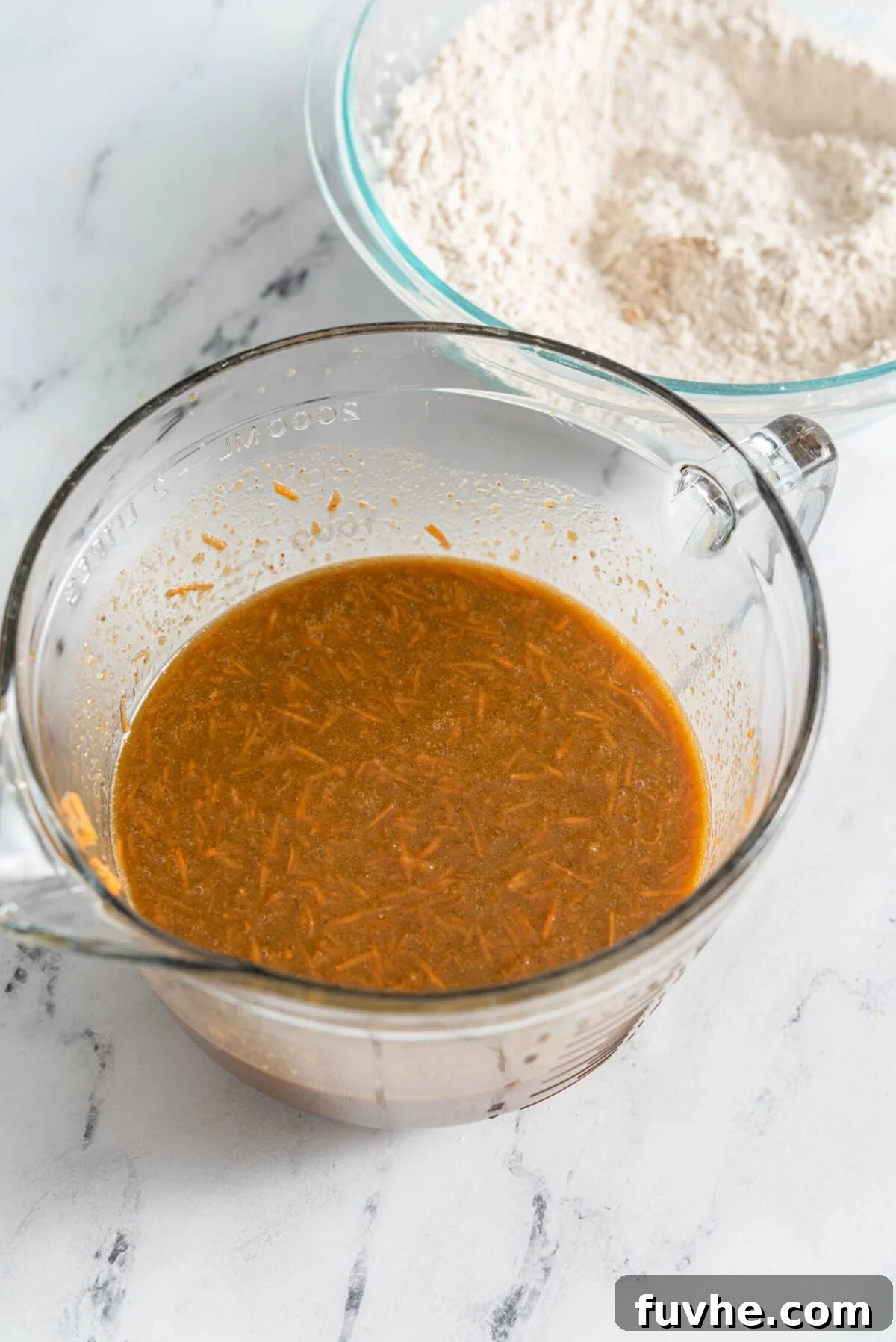 The Perfect Plant-Based Carrot Cake 5 A glass mixing bowl brimming with vibrant wet baking ingredients, including freshly grated carrots, all mixed together into a smooth, colorful batter.