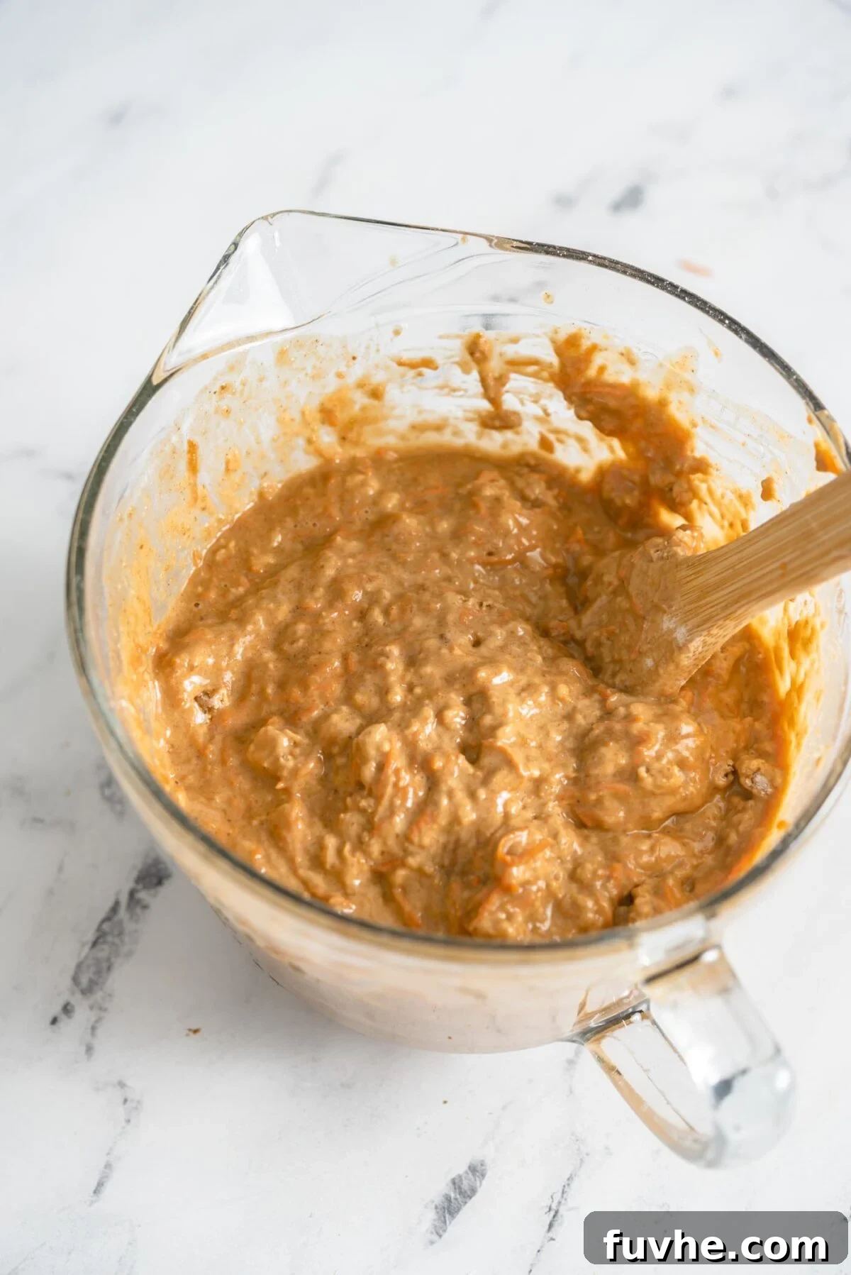 The Perfect Plant-Based Carrot Cake 6 A glass mixing bowl filled with raw vegan carrot cake batter, perfectly mixed with a wooden spoon, ready to be poured into baking pans.