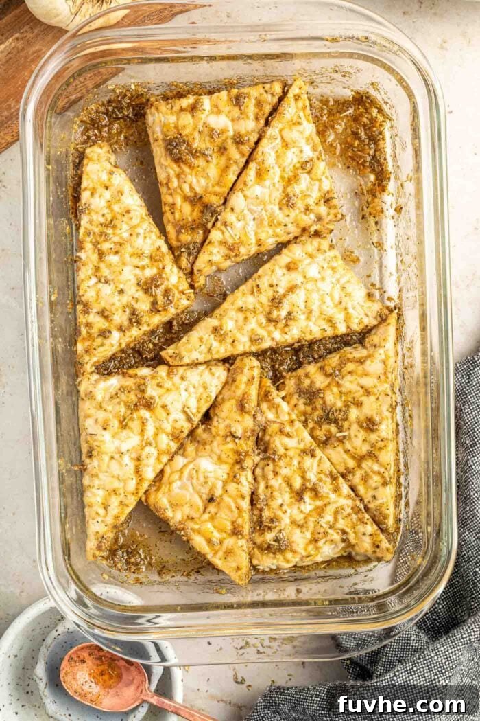 Thick, uniformly sliced tempeh pieces are being gently tossed in a rich herb and balsamic marinade within a glass food storage container, ensuring full flavor absorption.
