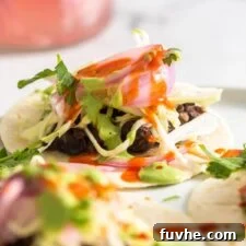 A small soft taco topped with pickled red onion, cabbage and cilantro crema on a plate. Two more tacos are blurred in the foreground.