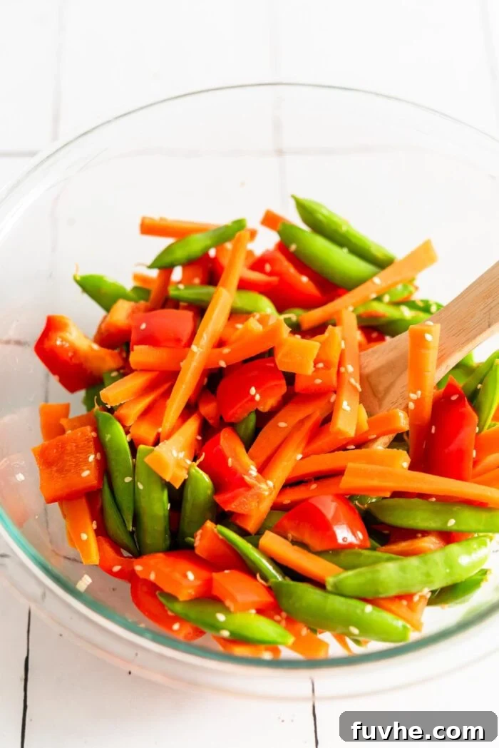 A glass mixing bowl holds a colorful medley of freshly stir-fried vegetables: bright green snap peas, vibrant orange matchstick carrots, and red bell pepper slices, lightly seasoned and ready to be added to a rice bowl.