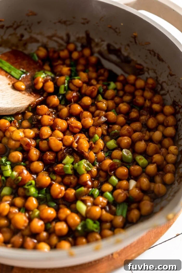 A close-up view of chickpeas simmering in a rich, glossy teriyaki sauce within a large skillet, garnished with bright green onions, showing the sauce perfectly thickening and coating the legumes.