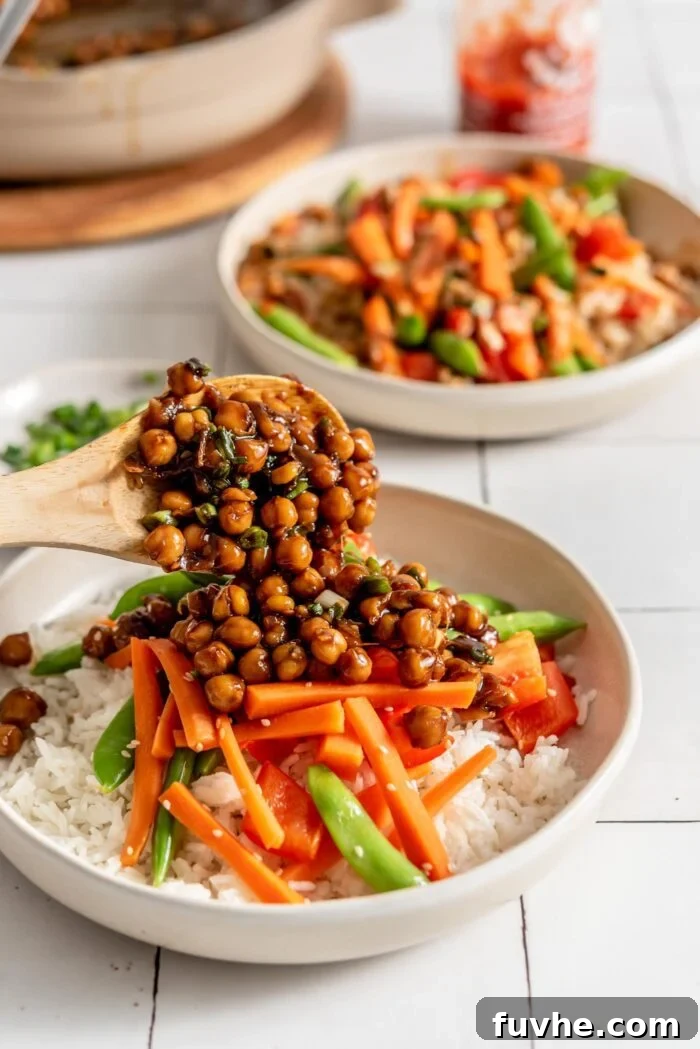 A hand holds a large spoon, scooping generous portions of teriyaki chickpeas over a bowl already filled with fluffy rice and colorful stir-fried vegetables. The bowl is set to be completed with sauce and garnishes, illustrating the final assembly stage of the dish.