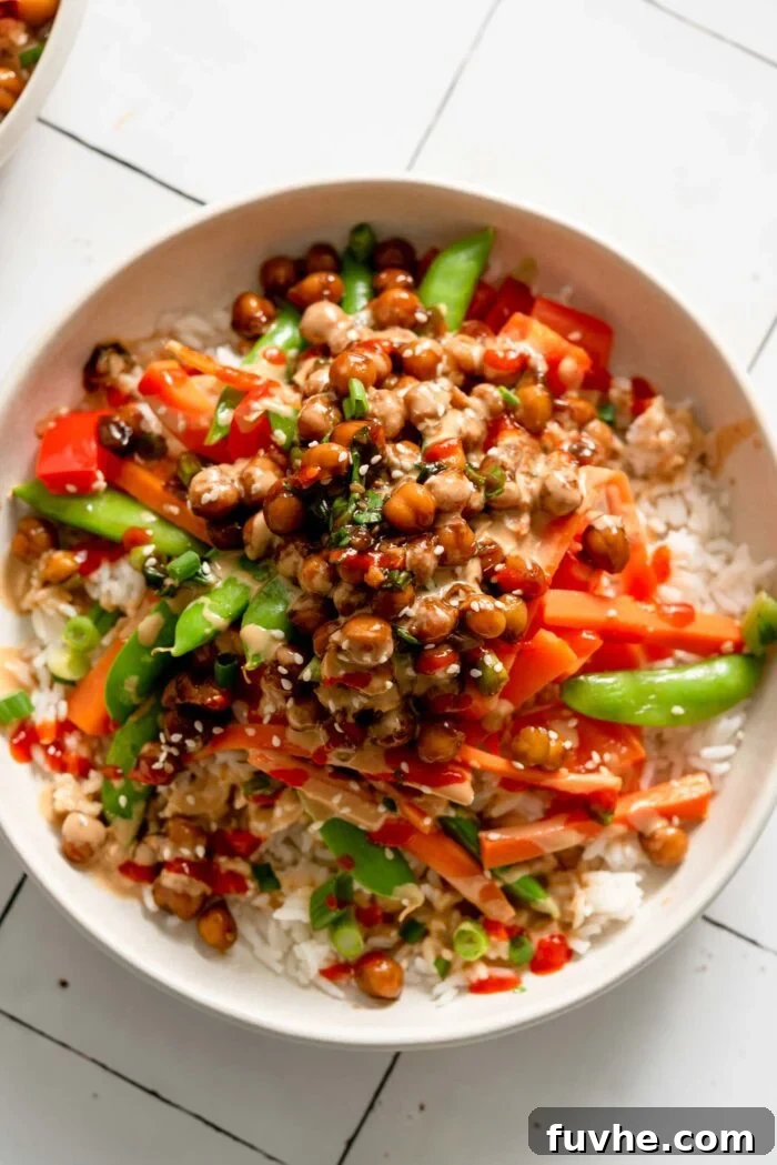 An overhead shot of a vibrant vegan Teriyaki Chickpea Rice Bowl, showcasing a harmonious arrangement of fluffy white rice, glistening teriyaki chickpeas, crisp snap peas, matchstick carrots, and red bell peppers. The dish is beautifully garnished with fresh green onions and sesame seeds, highlighting its colorful and appetizing presentation.