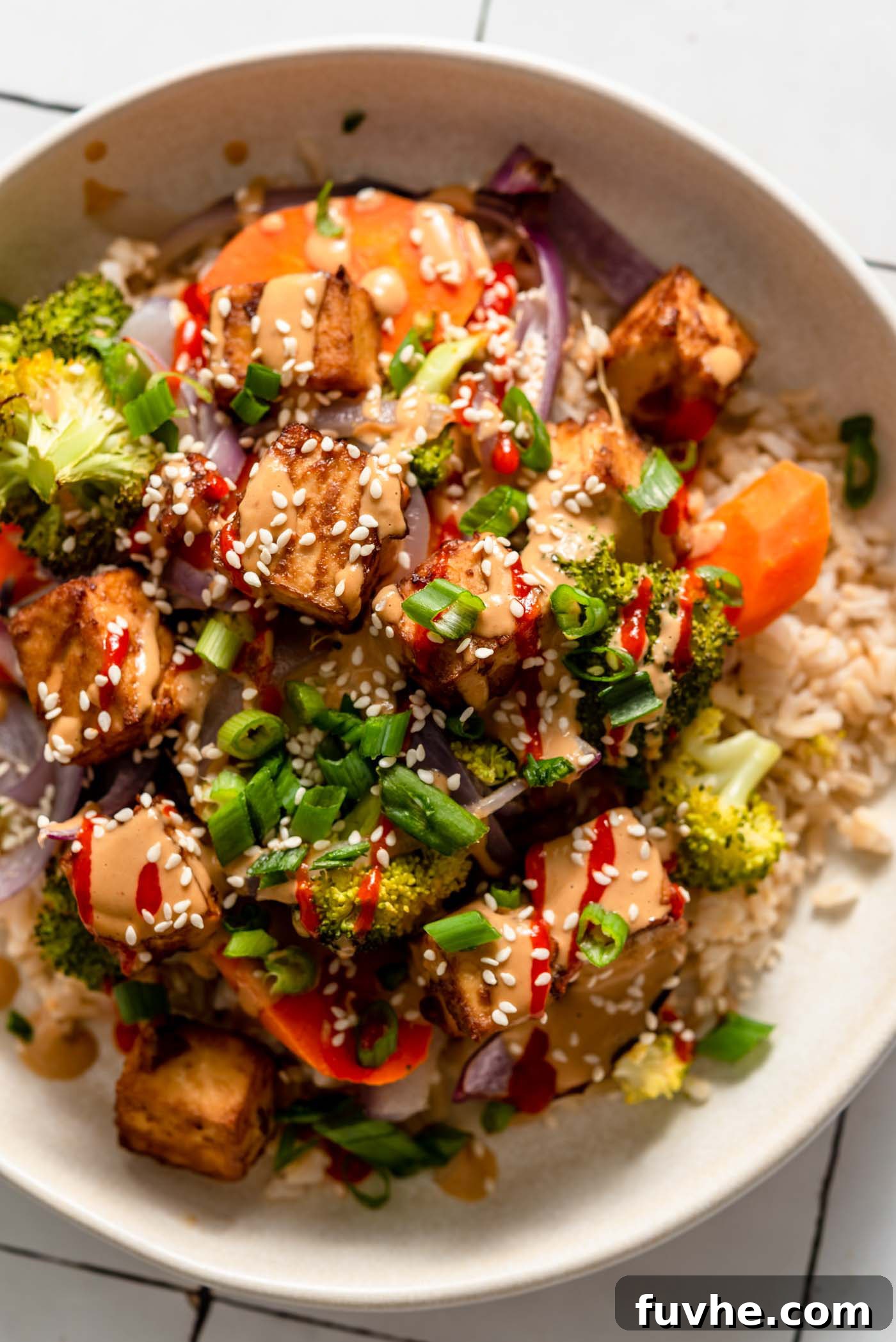 Overhead view of a colourful bowl with tofu, broccoli, carrot, onion and rice, topped with creamy peanut sauce, green onion, sesame seeds and hot sauce.
