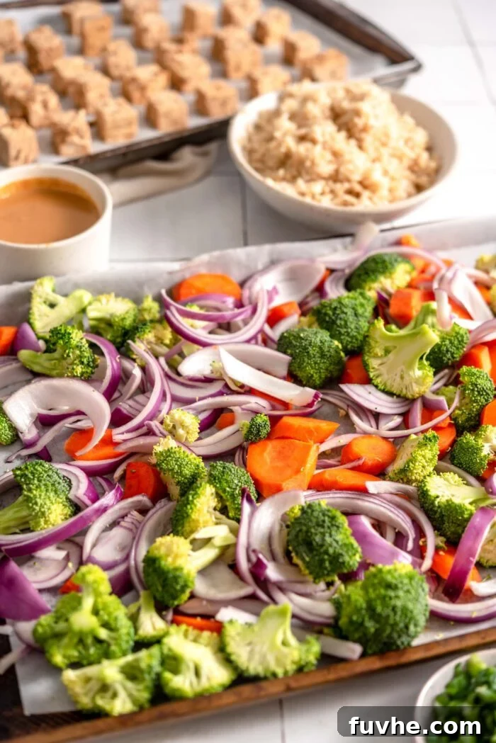 Chopped broccoli, onion and carrot on a baking pan. In the background, a pan with cubed tofu on it is visible.