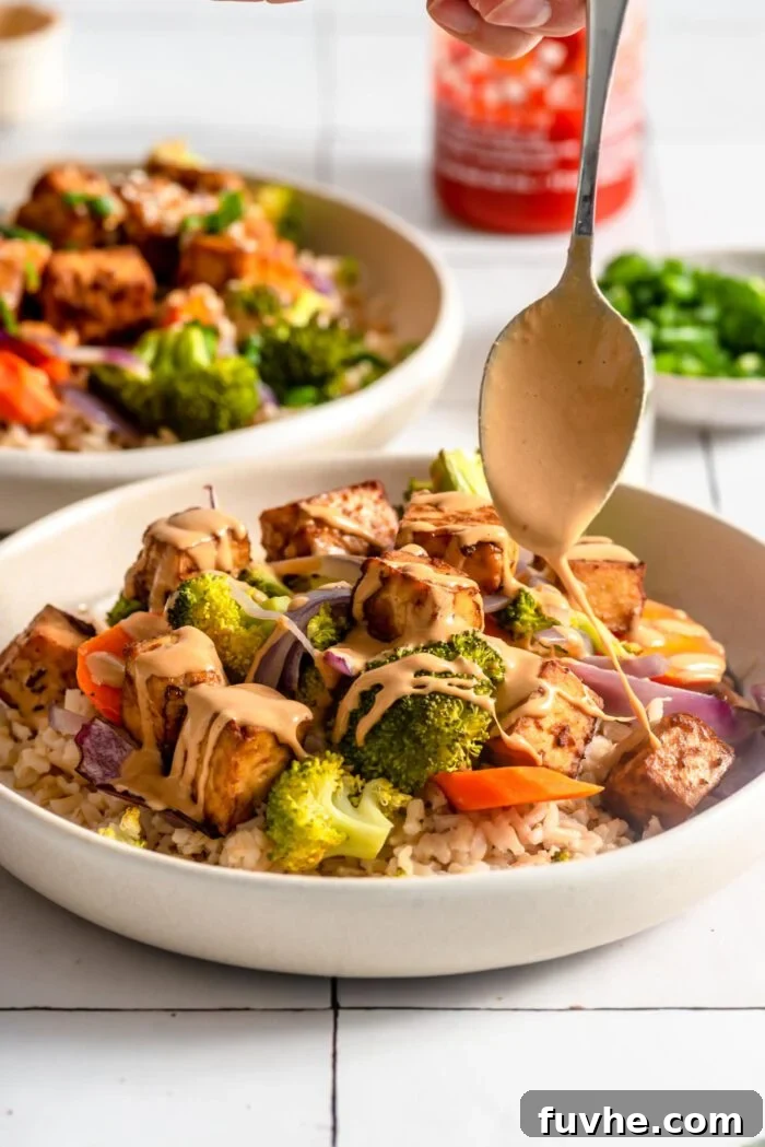 Spoon drizzling peanut sauce over a bowl of rice, vegetables and tofu.