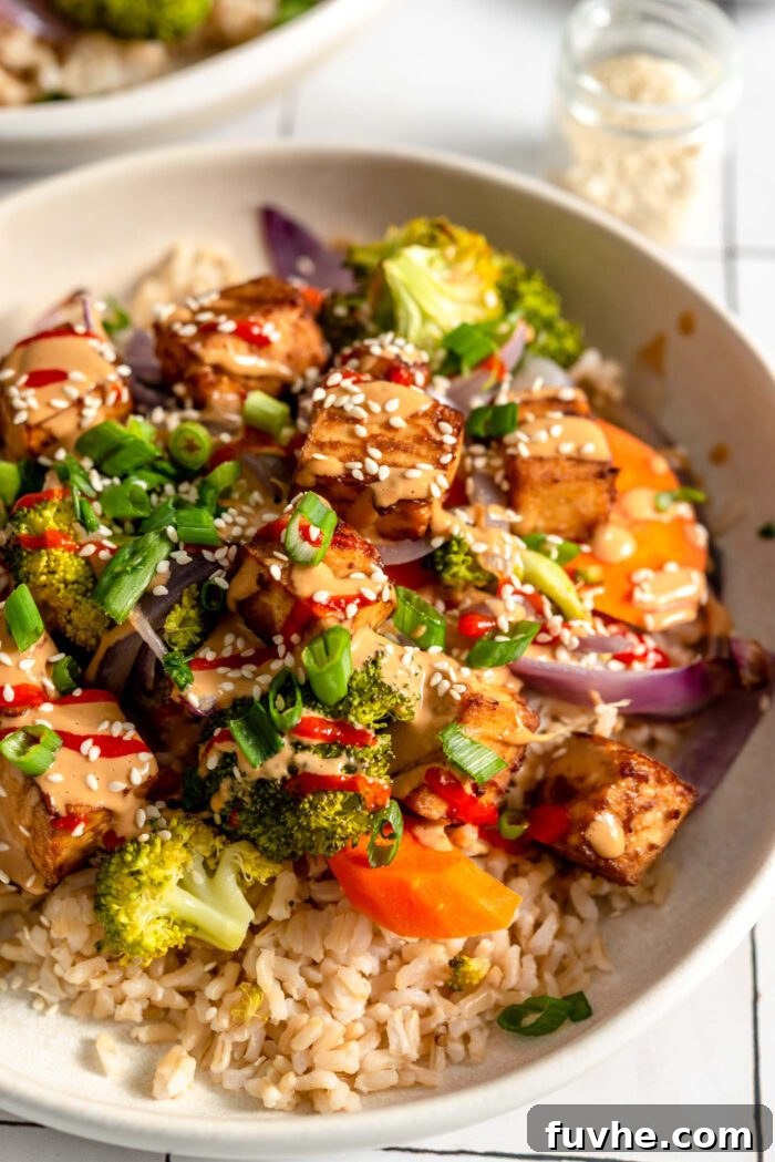 A colourful bowl with tofu, broccoli, carrot, onion and rice, topped with creamy peanut sauce, green onion, sesame seeds and hot sauce.