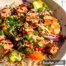 Overhead view of a colourful bowl with tofu, broccoli, carrot, onion and rice, topped with creamy peanut sauce, green onion, sesame seeds and hot sauce.