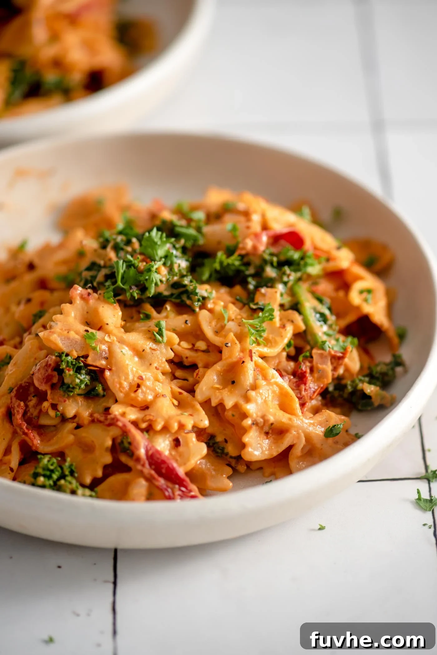 A bowl of creamy vegan sun-dried tomato pasta with fresh kale and parsley on top, highlighting its vibrant colors and rich texture.