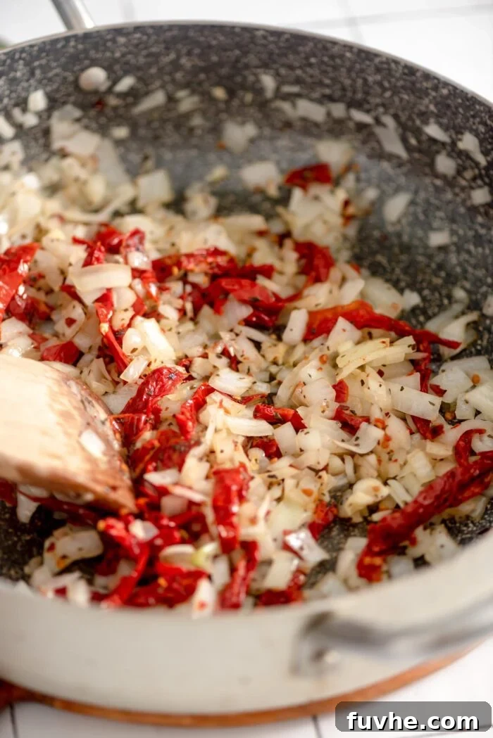 Finely diced onion, minced garlic, and chopped sun-dried tomatoes sizzling in a large skillet, releasing their aromatic flavors.