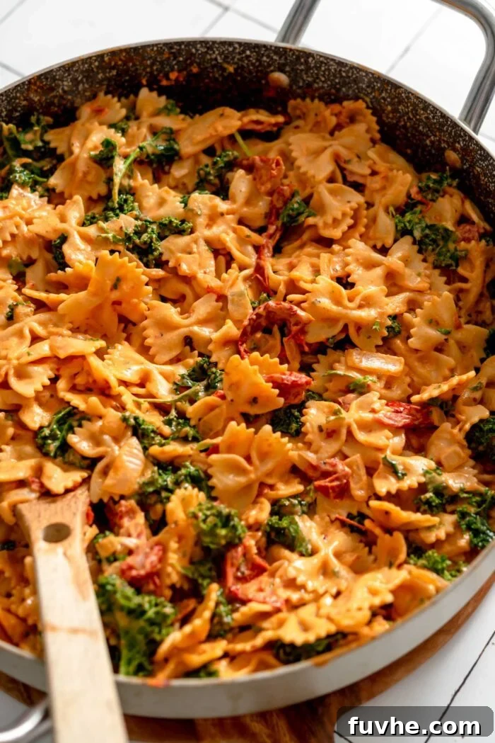 Creamy sun-dried tomato bowtie pasta with wilted kale in a large skillet, mixed with a wooden spoon, showcasing its perfect consistency.