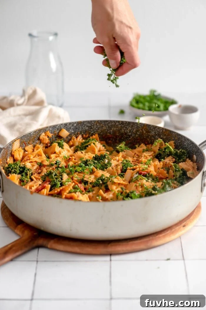 A hand gracefully sprinkling finely chopped fresh parsley over a large pan of creamy sun-dried tomato and kale pasta, adding a final touch of freshness.
