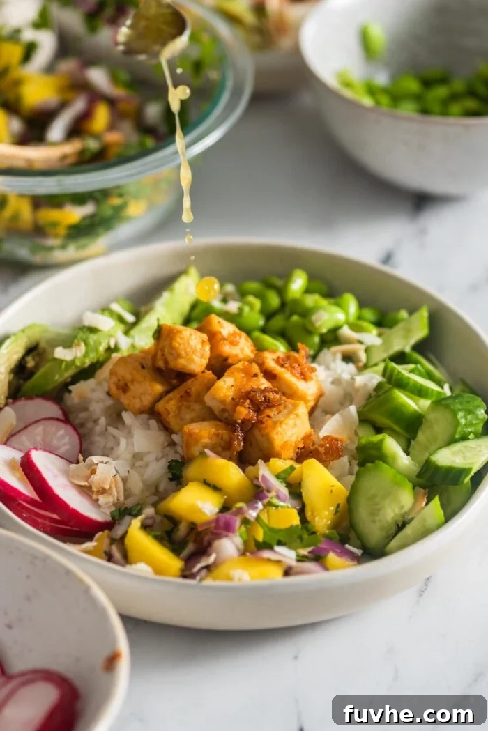 A small spoon drizzling ginger lime dressing over a colorful, fully assembled bowl with rice, mango salsa, edamame, avocado, and radish.
