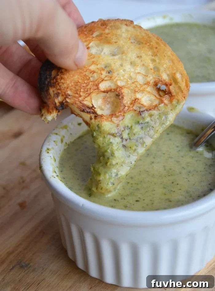 A hand dipping a crusty piece of sourdough bread into a small bowl of creamy vegan cheesy broccoli soup, highlighting its rich texture and appetizing appeal.