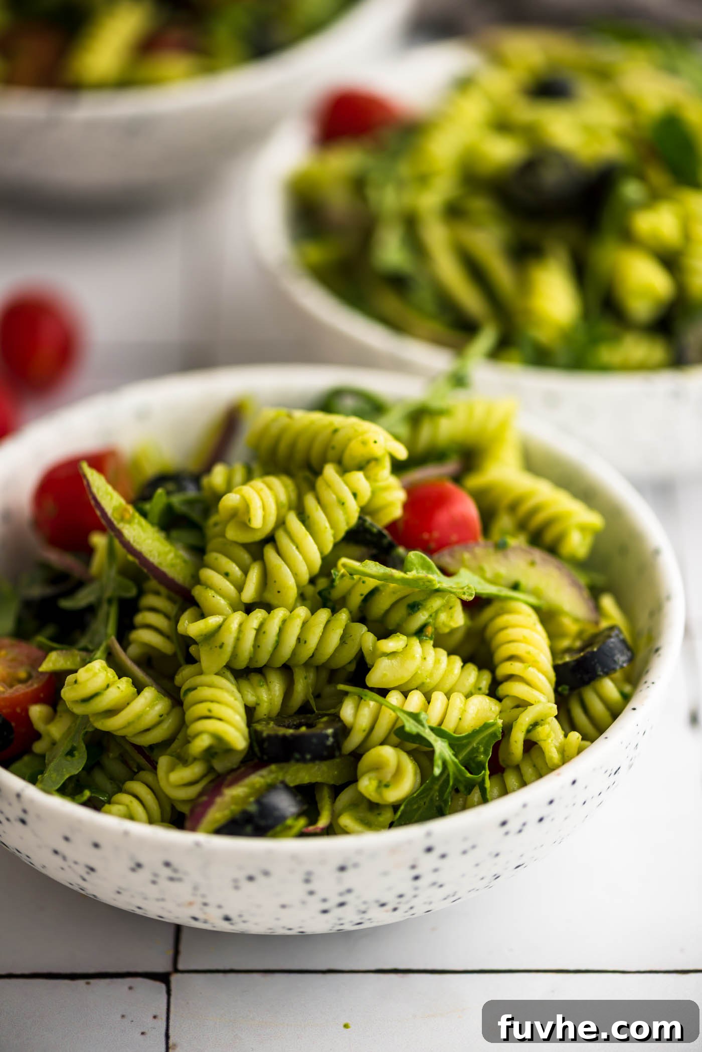 Vibrant Vegan Pesto Pasta Salad 2 A vibrant bowl of cold vegan pesto pasta salad, generously mixed with cherry tomatoes, fresh arugula, Kalamata olives, and thinly sliced red onion. This dish promises a refreshing and flavorful summer experience.