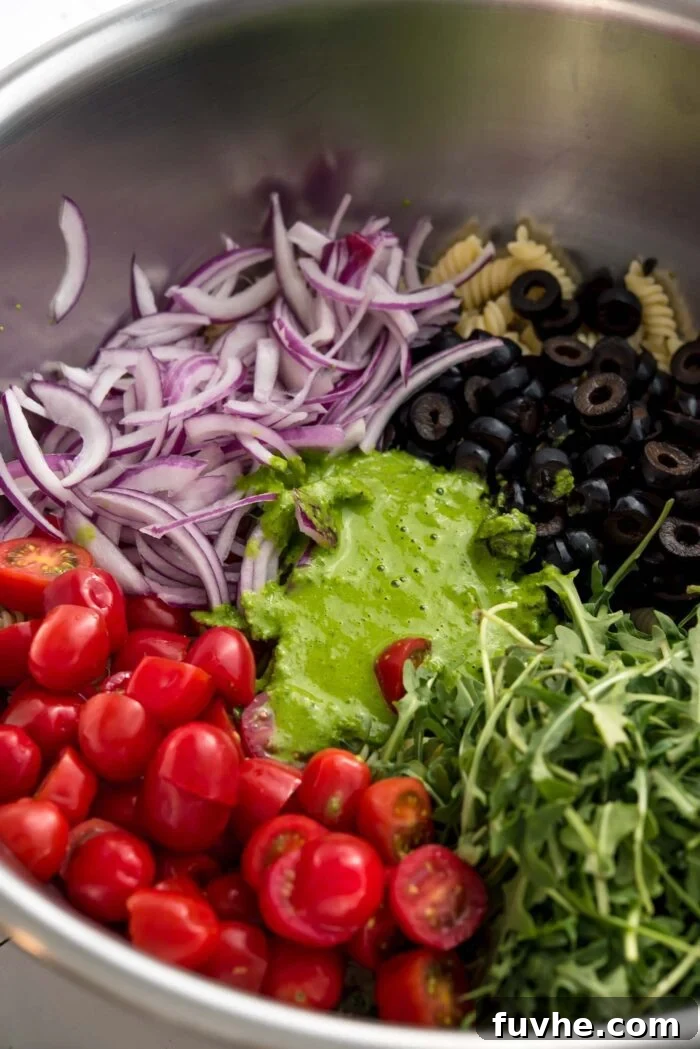 Vibrant Vegan Pesto Pasta Salad 5 A large metal mixing bowl filled with cooked pasta, halved cherry tomatoes, sliced black olives, fresh arugula, and thinly sliced red onion. A generous dollop of green pesto sauce is nestled on top, ready to be mixed into the vibrant salad ingredients.