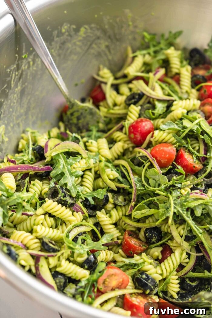 Vibrant Vegan Pesto Pasta Salad 6 A fork resting in a large metal mixing bowl, which holds a perfectly mixed cold pesto pasta salad. The salad is vibrant with pasta, tomatoes, arugula, and olives, highlighting its readiness to be served and enjoyed.