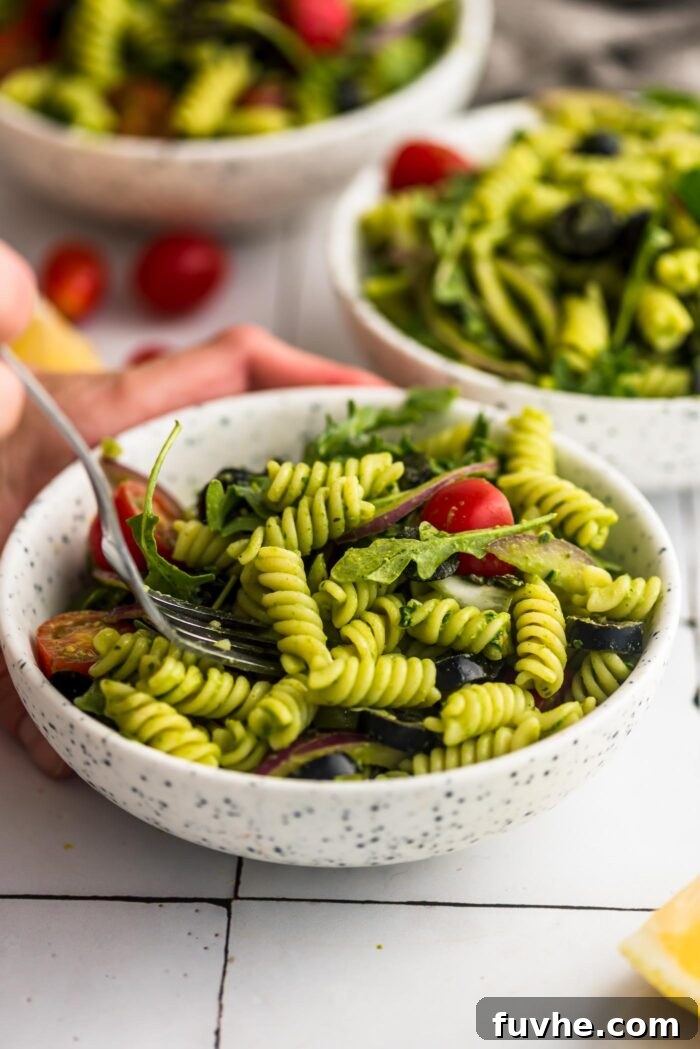 Vibrant Vegan Pesto Pasta Salad 7 A fork poised to scoop a portion of fresh pesto pasta salad from a small bowl. The salad is beautifully composed with pasta, halved cherry tomatoes, vibrant arugula, and black olives, ready for a delicious bite.