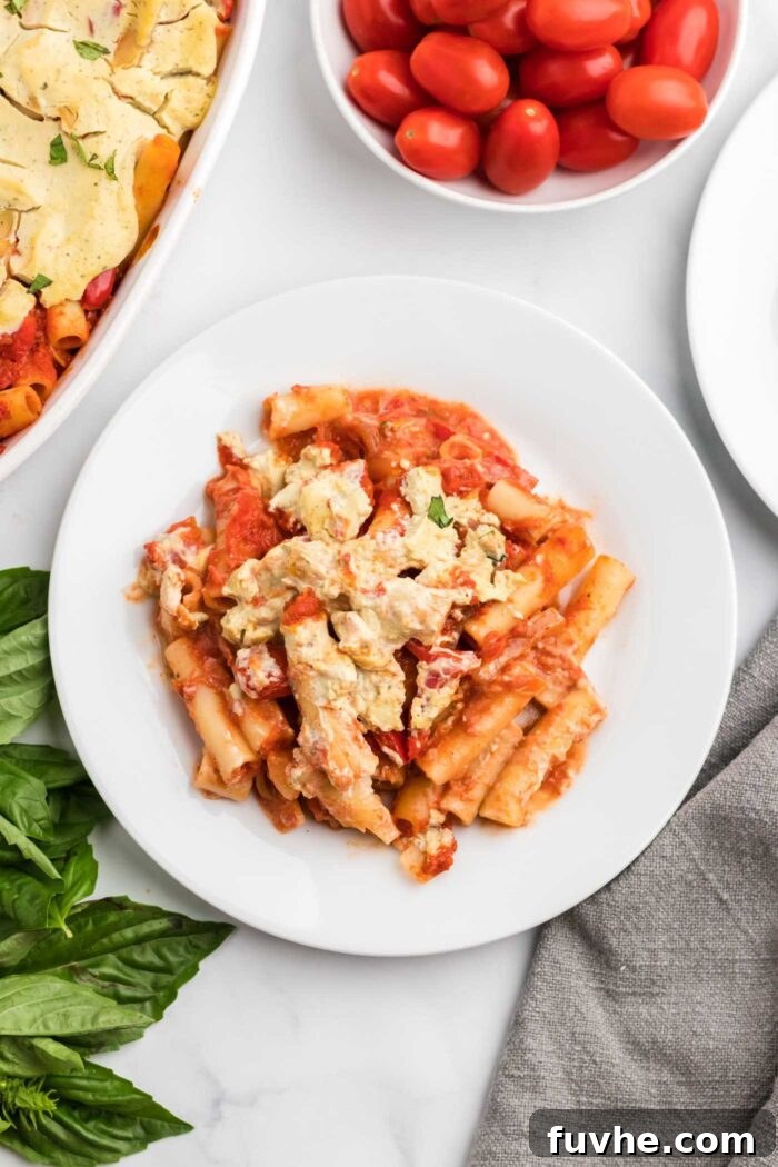 Hearty Plant-Based Baked Ziti 12 Overhead view of a serving of baked ziti made with tofu ricotta cheese on a small plate. There's a small dish or cherry tomatoes beside the plate and a baking dish of baked ziti.