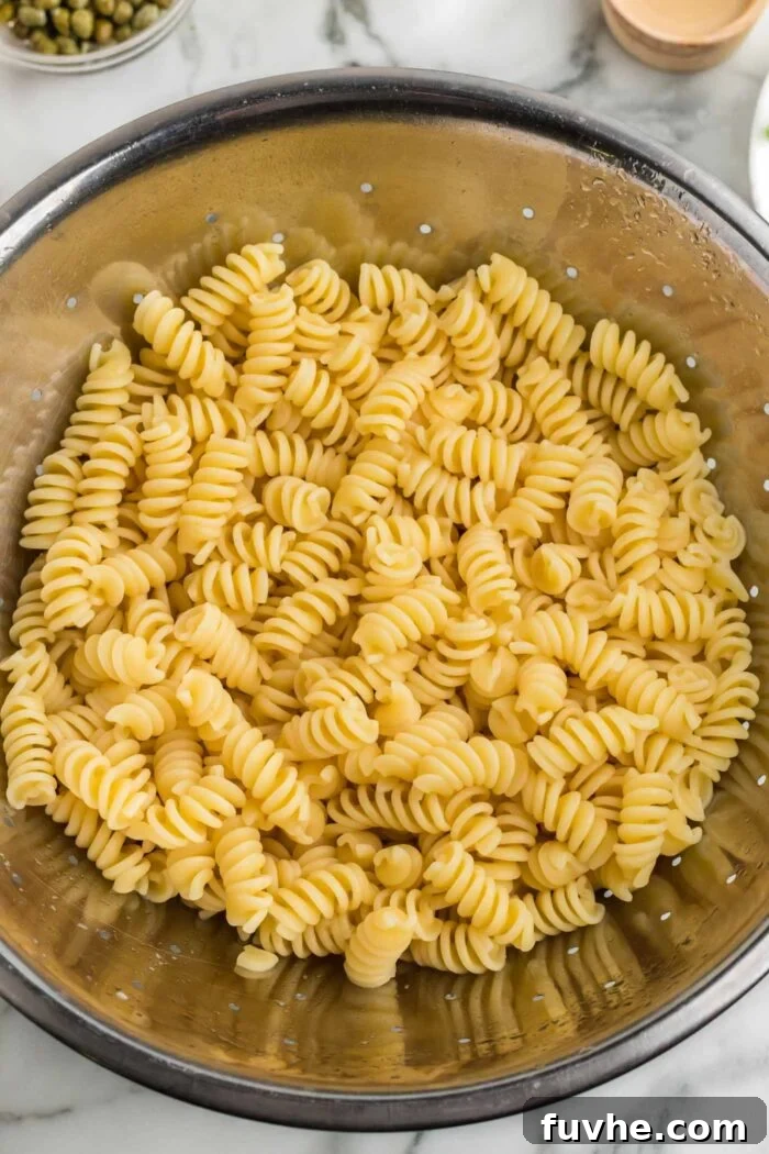 Freshly cooked rotini pasta noodles, perfectly al dente, resting in a gleaming metal colander after being rinsed under cold water. The steam has dissipated, indicating the pasta is ready for cooling and mixing into the salad.