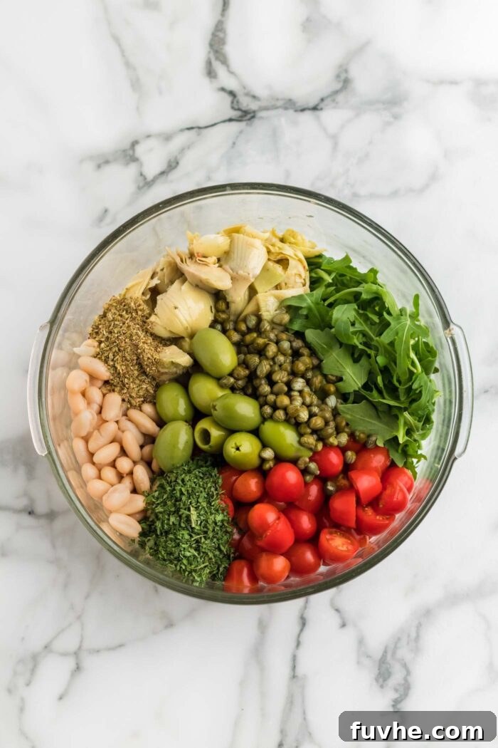 A vibrant collection of salad ingredients artfully arranged in a clear glass mixing bowl, awaiting the pasta. Visible are glistening green olives, bright red cherry tomatoes, fresh arugula, tender artichokes, tiny capers, aromatic spices, and creamy white kidney beans, ready to be tossed.