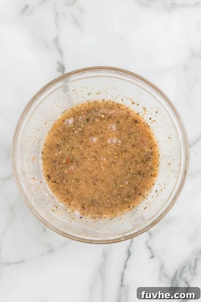 A small, clear glass bowl holding a vibrant, herb-infused vinaigrette, perfectly whisked and ready to dress the pasta salad. The light catches the subtle sheen of the olive oil and herbs, hinting at its zesty flavor.