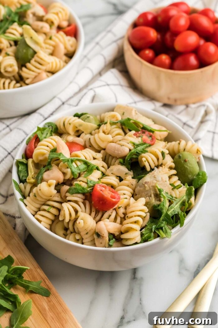 A refreshing bowl of zesty Italian pasta salad, brimming with colorful ingredients like diced tomatoes, fresh arugula, briny olives, and tender artichokes. A bowl of ripe tomatoes in the background reinforces the fresh appeal of the dish, ready for a delightful meal.