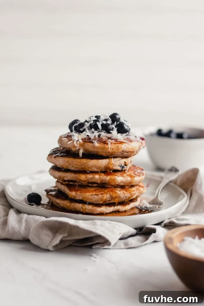 A tall stack of fluffy vegan blueberry coconut pancakes, generously topped with fresh blueberries, a sprinkle of shredded coconut, and a drizzle of maple syrup.