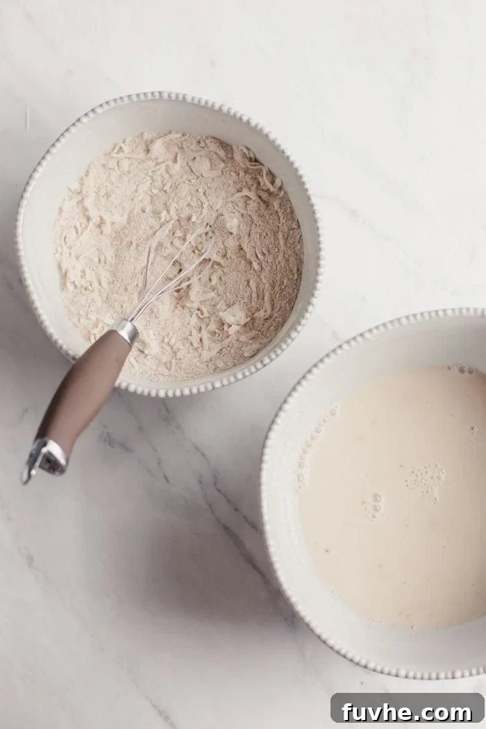 Two white bowls on a wooden surface: one containing dry ingredients like flour and shredded coconut, the other holding almond milk.