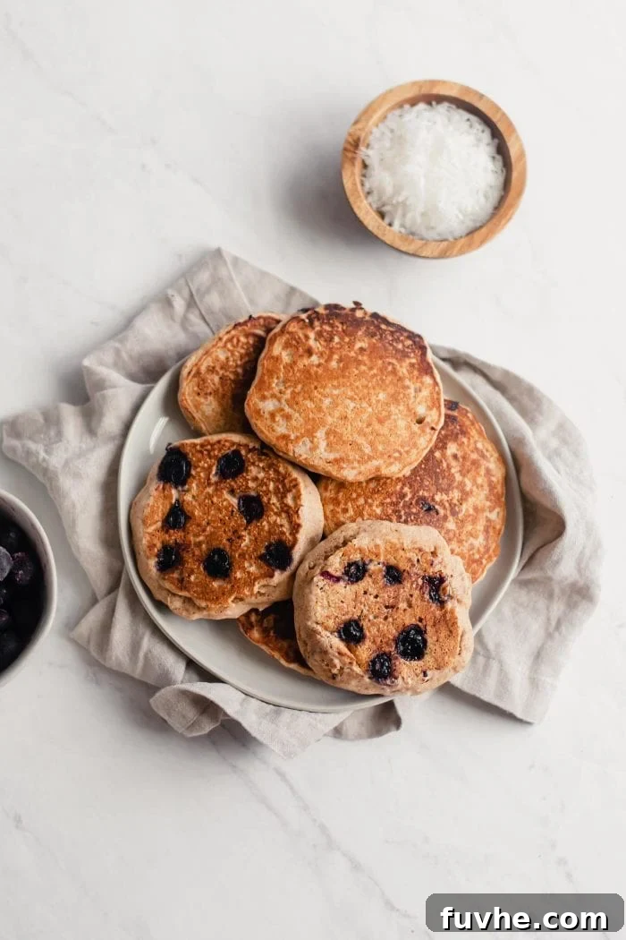 A beautifully arranged pile of golden brown blueberry coconut pancakes on a white plate, ready to be garnished.